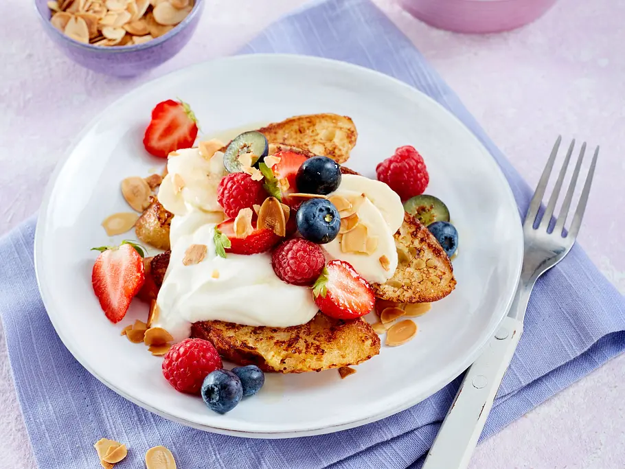  Drei goldbraune gebratene French Toast auf einem weißen Teller mit Quarkcreme, frischen Beeren und Mandelblättchen, daneben eine Gabel.