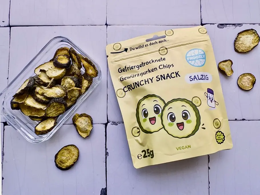 Auf einer hellen Fliesenfläche liegt eine Tüte von Froozly gefriergetrockneten Gewürzgurken-Chips, die ebenfalls in einer Glasschale daneben serviert sind.