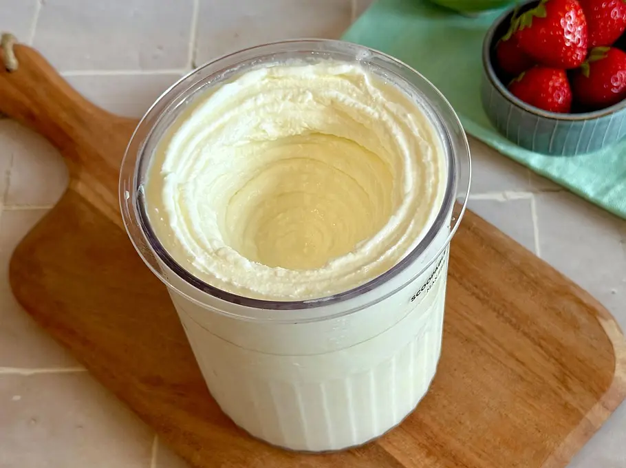 fertiger Frozen Joghurt im Zubereitungsbecher der Ninja Creami, platziert auf einem Holzbrett und im Hintergrund einige Früchte in Schalen.