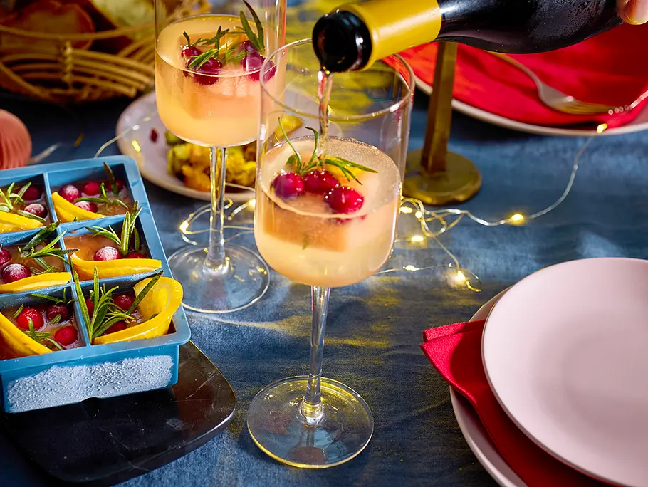 Frozen Weihnachts-Spritz Rezept Ein Weinglas, in dem ein Eiswürfel mit Cranberrys, Orange und Rosmarin liegt, wird mit Prosecco aufgefüllt