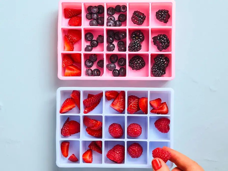 Eine Person verteilt verschiedene Beeren in die Mulden von zwei Silikoneiswürfelformen, eine rosa und eine hellblaue Form.