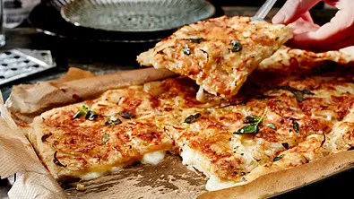 Eine Hand hebt ein Stück Fugazzeta mit geschmolzenem Käse und Basilikum von einem mit Backpapier ausgelegten Blech. - Foto: LECKER @ Bauer Media Group