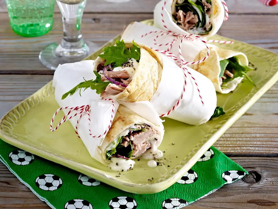 Vier halbierte Wraps mit Thunfisch, Salat und Dressing, in Papier gewickelt und mit roter Kordel gebunden, serviert auf einer grünen Platte. Daneben stehen zwei Gläser und eine Serviette mit Fußballmotiv.