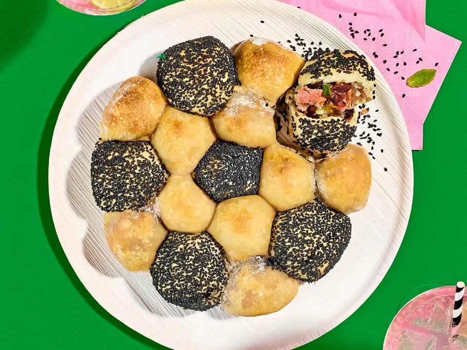 WM-Zupfbrot mit Salami-Käse-Füllung Rezept Zupfbrot-Bällchen mit Salami-Käse-Füllung, teils mit Mohn oder Sesam bestreut, angerichtet auf einem weißen Teller.