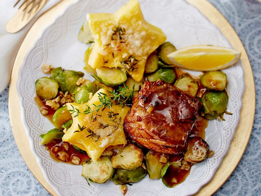 Ein Gänsebrust-Tournedo mit glänzender Bratensauce, serviert auf einem weißen Teller mit goldener Umrandung, dazu gebratene Rosenkohlhälften, zwei lockere Kartoffelkissen mit Kräutern und ein Zitronenschnitz.