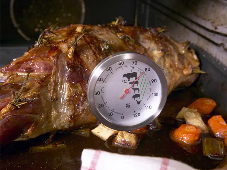 Eine gebratene Lammkeule mit goldbrauner Kruste liegt in einer Bratform, ein Bratenthermometer misst die Kerntemperatur, ringsum geröstetes Wurzelgemüse.