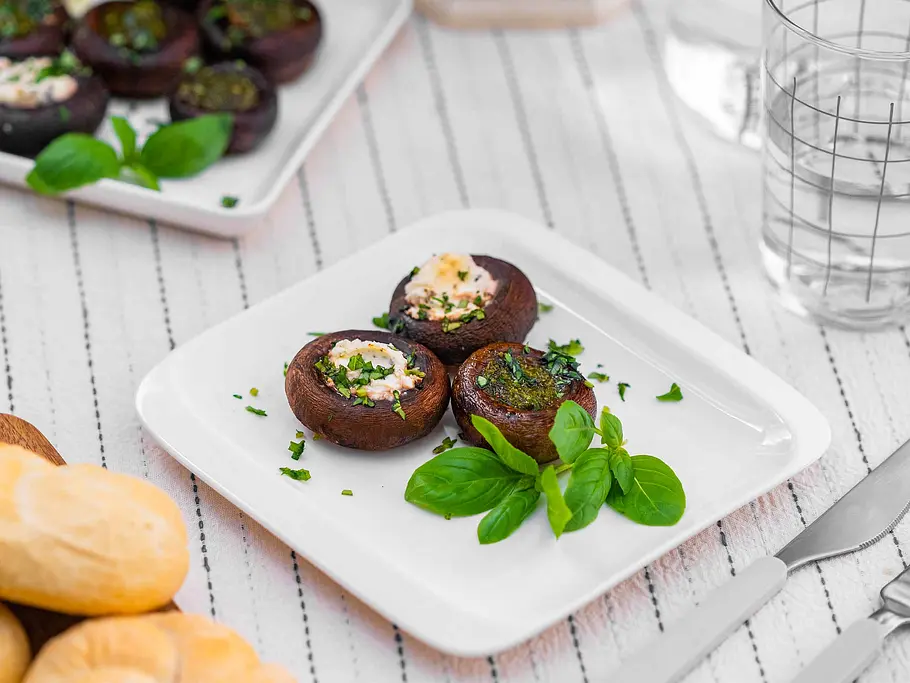 Drei mit Kräuter-Frischkäse und grünem Pesto gefüllte Champignons liegen auf einem weißen Teller, daneben frisches Basilikum als Garnitur.