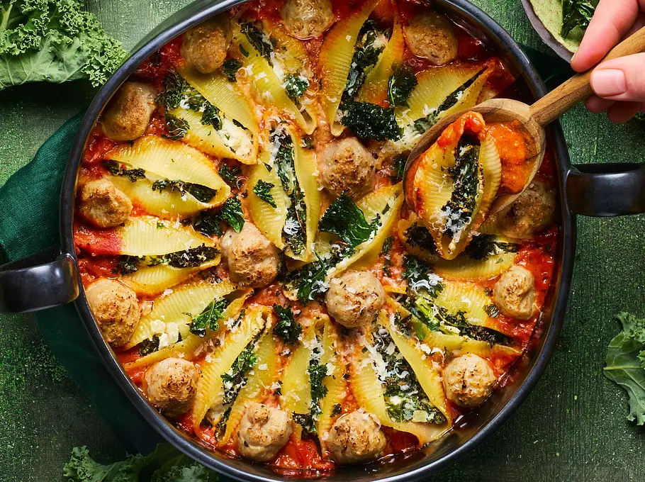 Ofenform mit großen Muschelnudeln, gefüllt mit Grünkohl und Käse, dazu rote Tomatensauce und goldene Käsekruste, auf grünem Untergrund mit passendem grünen Teller daneben.