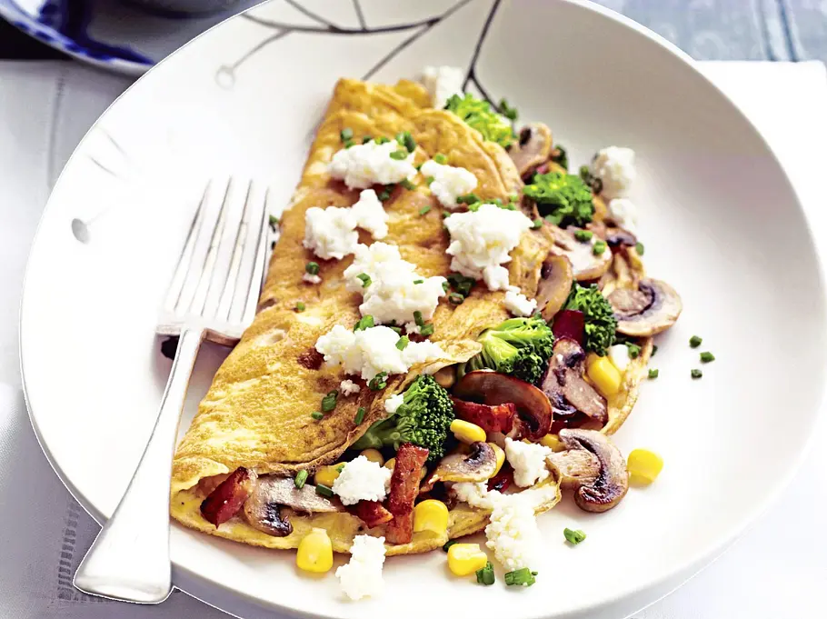 Gefülltes Omelett mit Brokkoli und Pilzen Rezept Omelett gefüllt mit Brokkoli, Pilzen, Mais und Tomaten, bestreut mit Frischkäse, angerichtet auf einem weißen Teller mit einer Gabel, daneben eine Schale Rucola.