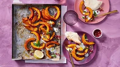 Eine Grillschale mit mehreren gegrillten Kürbisspalten, die mit Granatapfelkernen, Zitrone und Kräutern garniert sind, steht auf einem violetten Untergrund neben zwei Tellern, auf denen bereits ein paar gegrillte Hokkaido-Spalten mit Fladenbrotstücken liegen. - Foto: LECKER @ Bauer Media Group