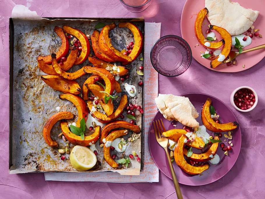 Eine Grillschale mit mehreren gegrillten Kürbisspalten, die mit Granatapfelkernen, Zitrone und Kräutern garniert sind, steht auf einem violetten Untergrund neben zwei Tellern, auf denen bereits ein paar gegrillte Hokkaido-Spalten mit Fladenbrotstücken liegen.