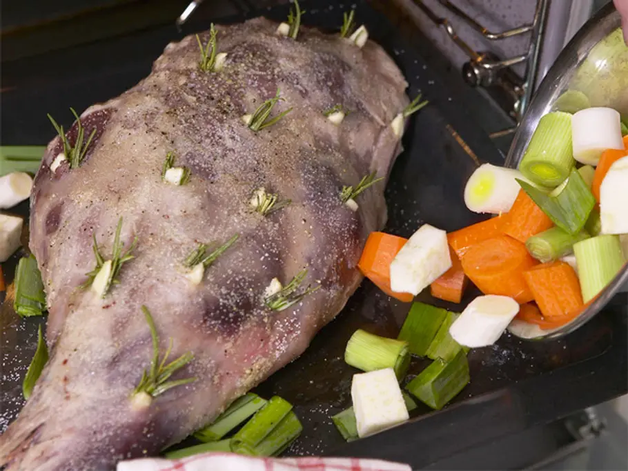 Eine gewürzte, mit Rosmarin und Knoblauch gespickte Lammkeule liegt in einer Bratform; daneben werden grob geschnittene Möhren, Sellerie und Lauch in die Form gegeben.