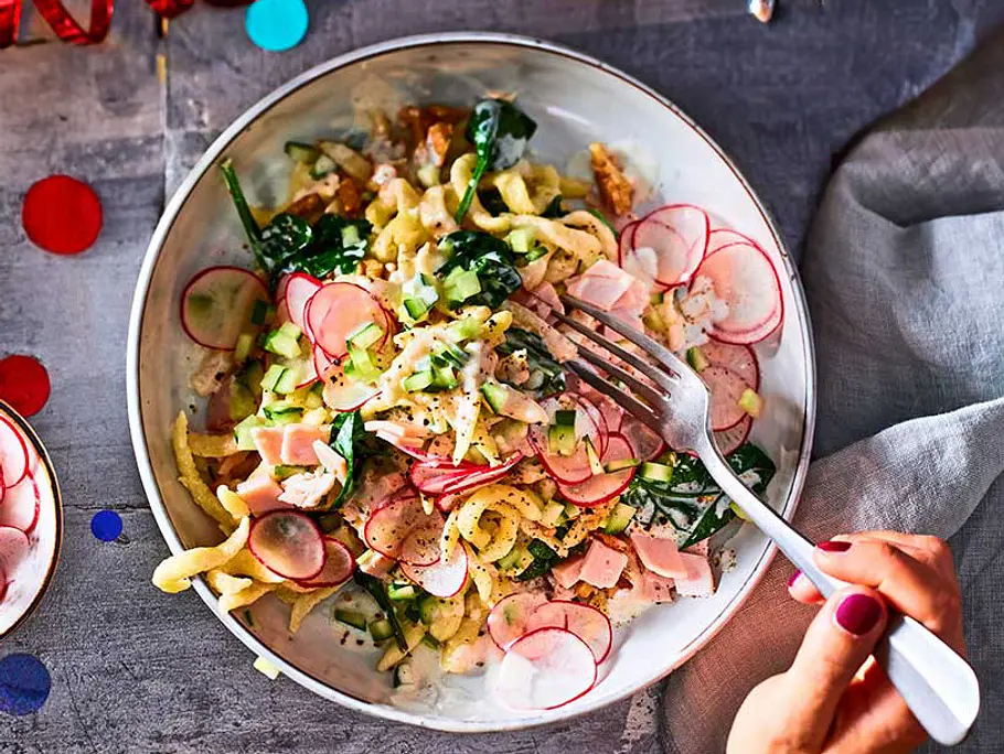 Spätzle-Salat mit Radieschenscheiben, Gurke, Spinat und Schinken in cremigem Joghurt-Dressing, serviert in einer Schüssel mit Gabel.