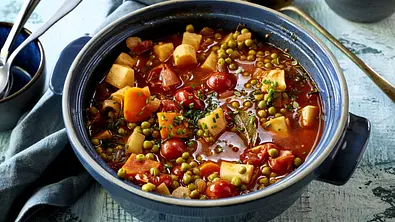 Bunter Gemüseeintopf mit Erbsen, Tomaten, Karotten und weiteren Gemüsewürfeln in aromatischer Brühe, serviert in einem rustikalen blauen Topf auf Holztisch mit Servierlöffel daneben. - Foto: LECKER @ Bauer Media Group