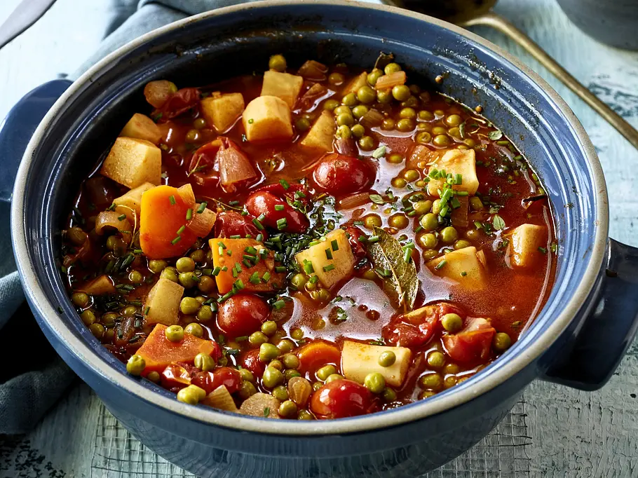 Bunter Gemüseeintopf mit Erbsen, Tomaten, Karotten und weiteren Gemüsewürfeln in aromatischer Brühe, serviert in einem rustikalen blauen Topf auf Holztisch mit Servierlöffel daneben.