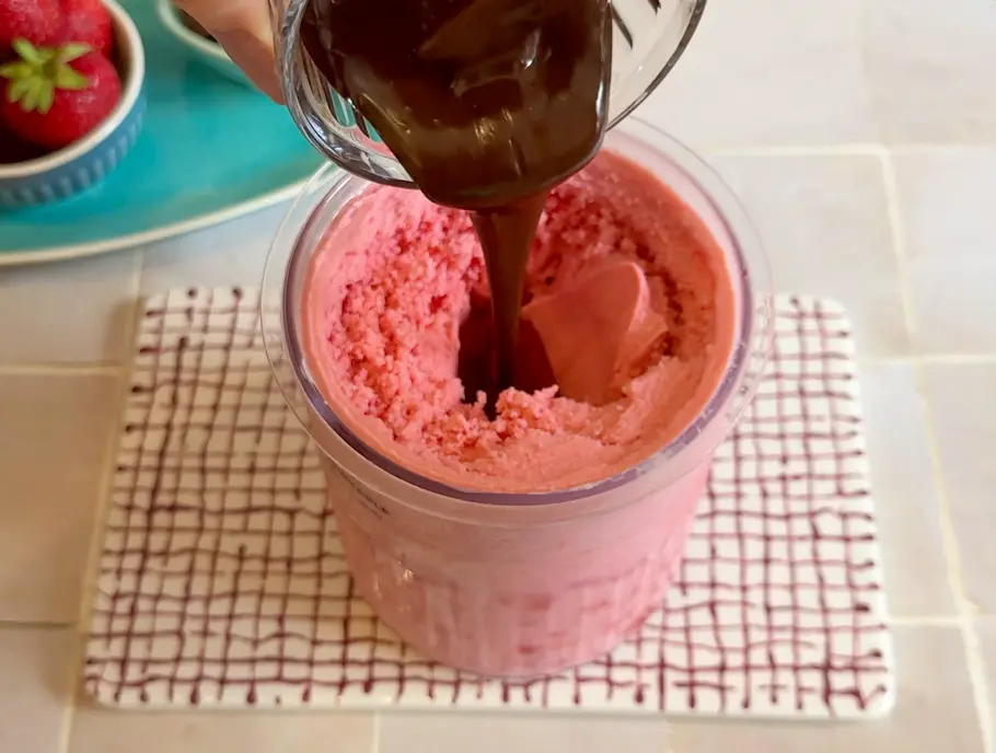 Geschmolzene dunkle Schokolade wird aus einem kleinen Gefäß in den Erdbeer-Frozen-Joghurt gegossen 