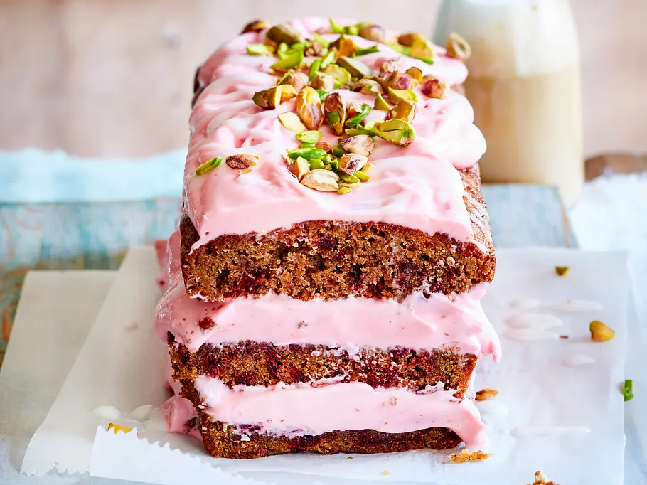 Kastenkuchen mit rosa Frischkäsecreme und Pistazien bestreut, angeschnitten und aufeinander gestapelt auf Backpapier.