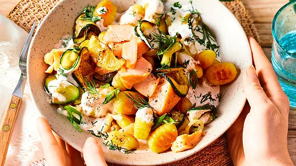 Gnocchi-Pfanne mit Zucchini, Lauchzwiebeln, Lachs und Dill in einer Schale, die von zwei Händen umfasst wird - Foto: LECKER @ Bauer Media Group
