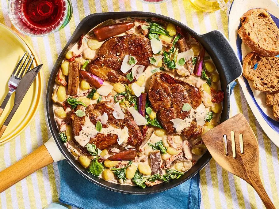 Gnocchi-Spinat-Pfanne mit zart gebratenen Steakstücken in einer cremigen Sauce, serviert in einer Pfanne auf einem gelb-weiß gestreiften Tischtuch, dazu Brotscheiben auf einem kleinen Teller und zwei Gläser mit rotem Getränk.