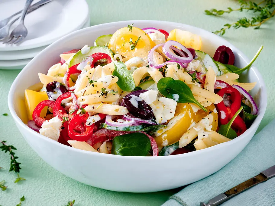Griechischer Nudelsalat mit Feta-Dressing Eine weiße Schüssel mit cremigen Nudelsalat mit Oliven, Feta-Dressing, roten Zwiebeln und Salatblättern.