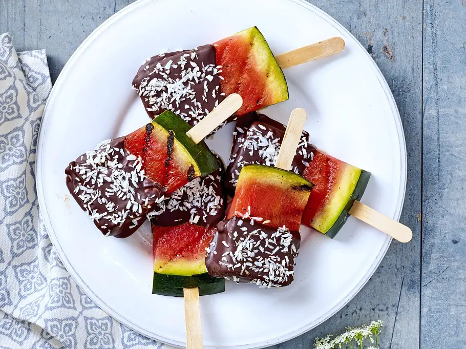 Gegrillte Melonen-Popsicles Rezept Wassermelonenstücke am Stiel, teilweise mit Schokoladenguss überzogen und mit Kokosraspeln bestreut, auf einem weißen Teller arrangiert.