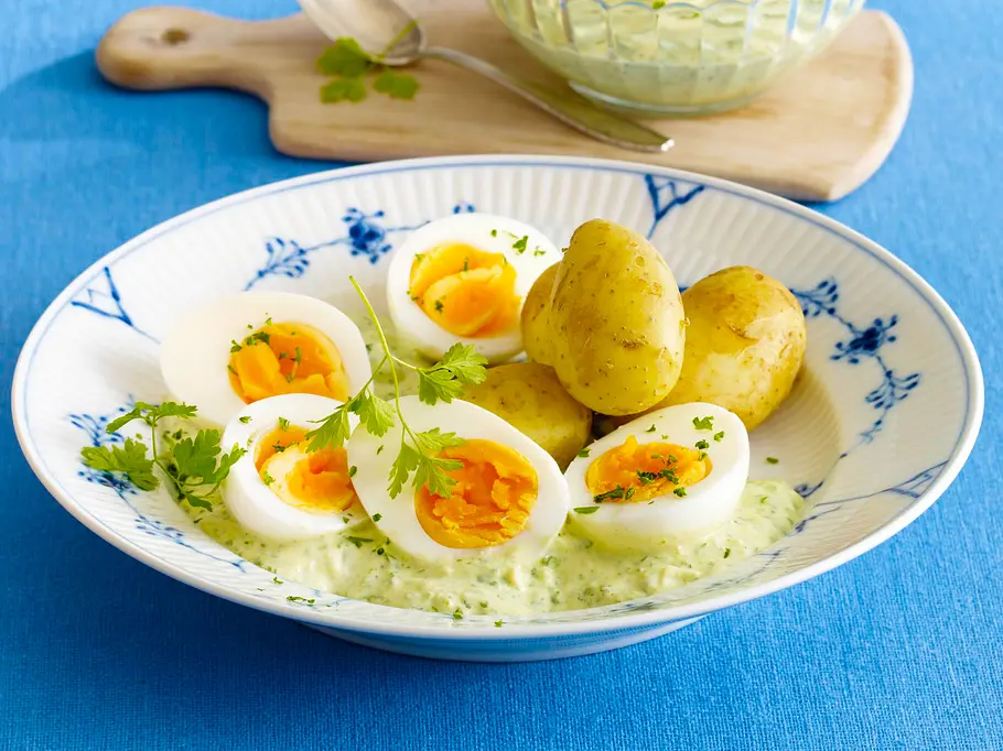 Grüne Soße mit neuen Kartoffeln Rezept Gekochte Eierhälften und Pellkartoffeln mit cremiger Kräutersoße auf einem blauen Teller, garniert mit frischen Kräutern.