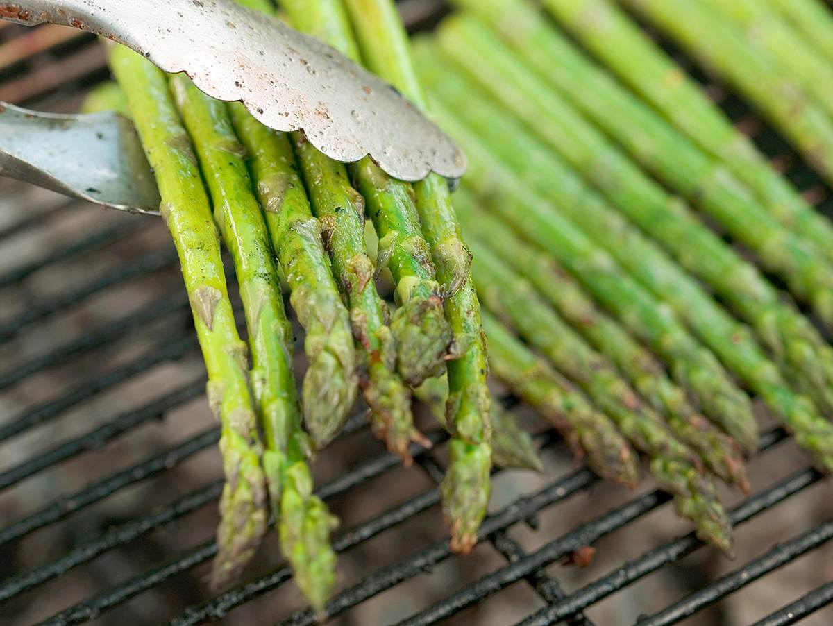 Grüner Spargel liegt auf dem Grill und wird mit einer Grillzange gewendet