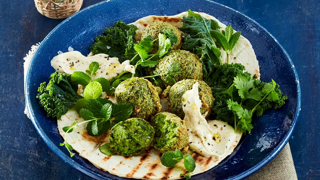 Knusprige, grüne Grünkohl-Falafel auf einem Fladenbrot, serviert mit cremigem Dip, frischen Kräutern und Grünkohlblättern auf einem dunkelblauen Teller. - Foto: LECKER @ Bauer Media Group Knusprige, grüne Grünkohl-Falafel auf einem Fladenbrot, serviert mit cremigem Dip, frischen Kräutern und Grünkohlblättern auf einem dunkelblauen Teller. - Foto: LECKER @ Bauer Media Group