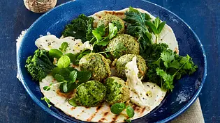 Knusprige, grüne Grünkohl-Falafel auf einem Fladenbrot, serviert mit cremigem Dip, frischen Kräutern und Grünkohlblättern auf einem dunkelblauen Teller. - Foto: LECKER @ Bauer Media Group