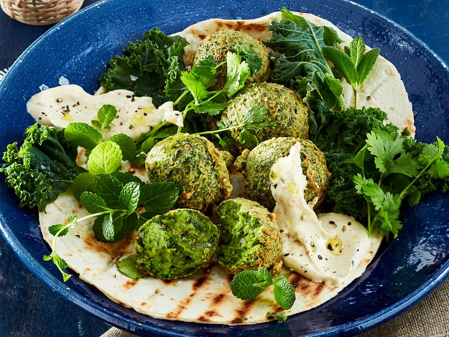 Knusprige, grüne Grünkohl-Falafel auf einem Fladenbrot, serviert mit cremigem Dip, frischen Kräutern und Grünkohlblättern auf einem dunkelblauen Teller.