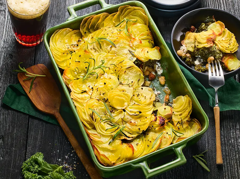 Grünkohl-Kartoffel-Gratin mit goldbraun überbackenen, dünn geschnittenen Kartoffelscheiben in einer grünen Auflaufform, serviert mit frischen Kräutern auf dunklem Holztisch, daneben schwarze Schale mit einer Portion Gratin und ein Glas Bier.