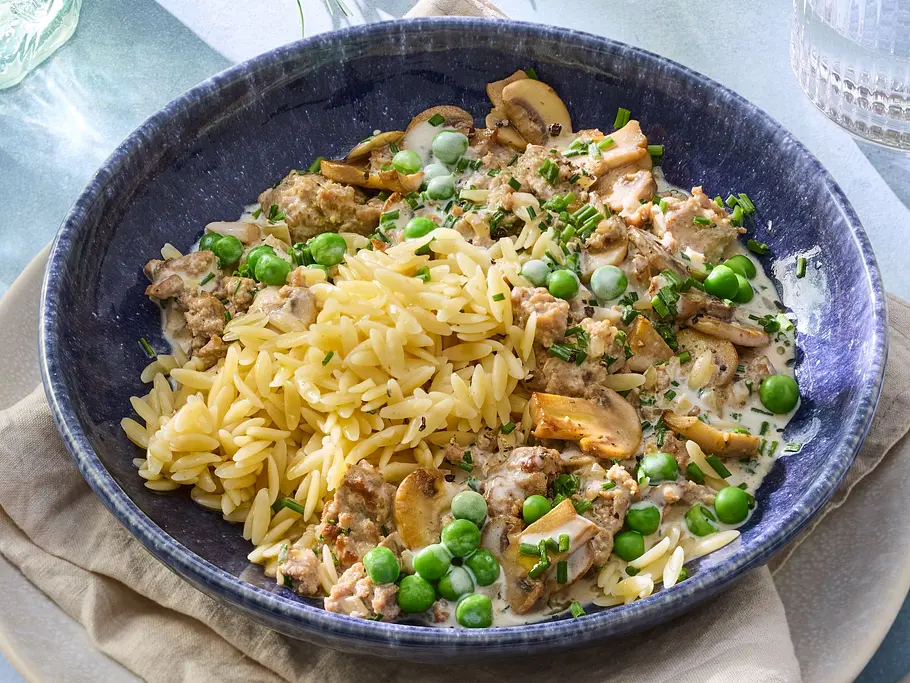 Pilzrahm mit Kritharaki Rezept Kritharaki-Nudeln in einem tiefen blauen Teller, serviert mit cremigem Pilzrahm, frischen Erbsen und fein gehacktem Schnittlauch. Im Hintergrund eine Pfanne mit Pilzrahm auf einem Holzbrett und Kräutern.
