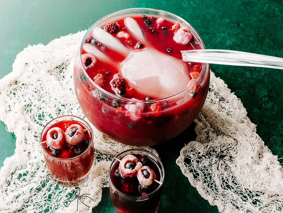 Halloween-Bowle Rezept Rote Bowle mit Beeren, großen Eishand und Fruchtaugen in Gläsern, dekoriert mit hellem Tuch und Kunststoffspinnen.