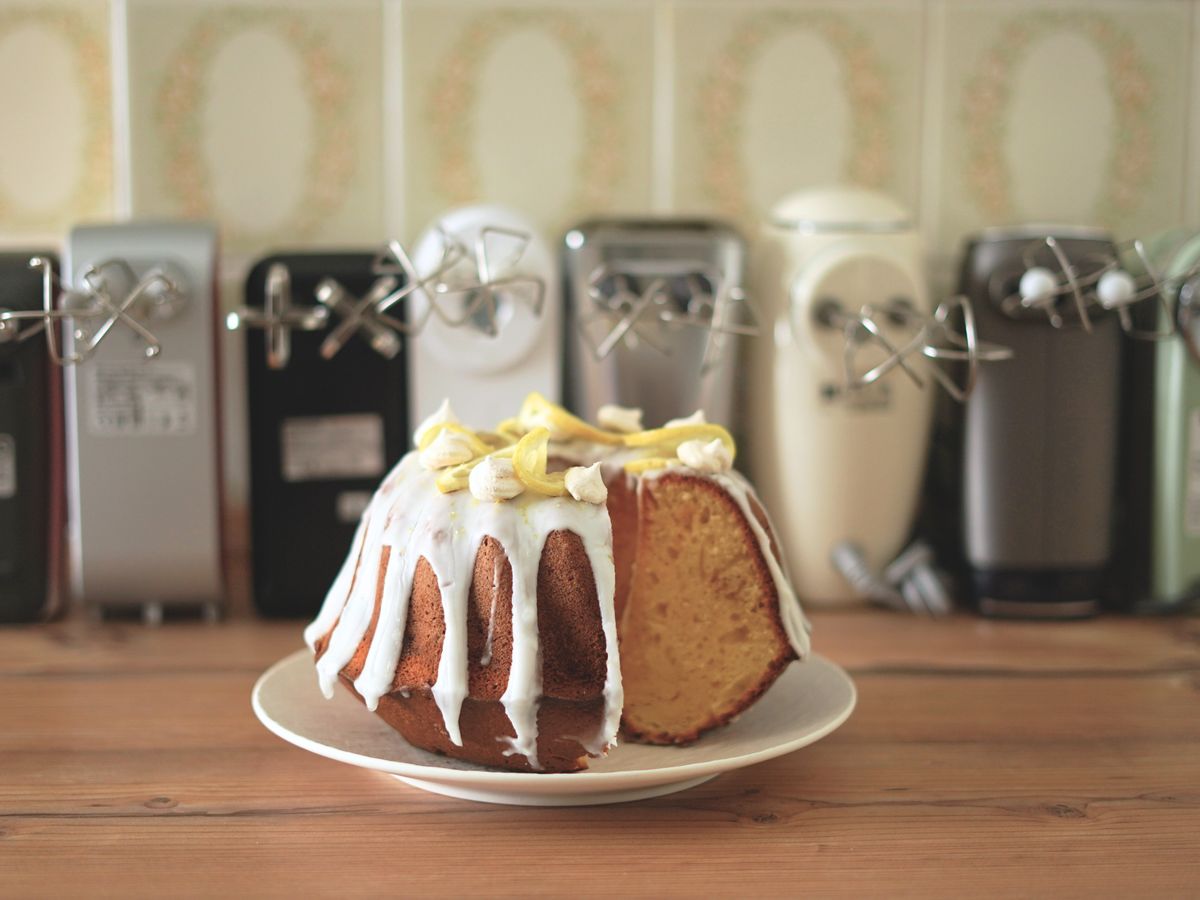 Ein angeschnittener Zitronen-Gugelhupf-Rührkuchen steht auf einer hölzernen Arbeitsplatte vor einer Reihe Handmixer mit Quirlen