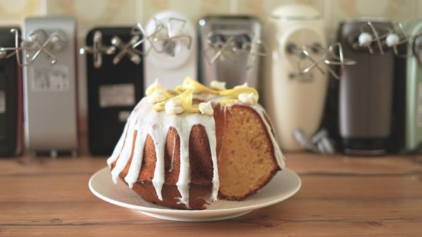 Ein angeschnittener Zitronen-Gugelhupf-Rührkuchen steht auf einer hölzernen Arbeitsplatte vor einer Reihe Handmixer mit Quirlen - Foto: LECKER.de