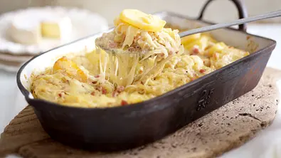 Ein Löffel hebt eine Portion Kartoffelauflauf aus einer dunklen Auflaufform. Geschmolzener Käse zieht lange Fäden. - Foto: LECKER @ Bauer Media Group