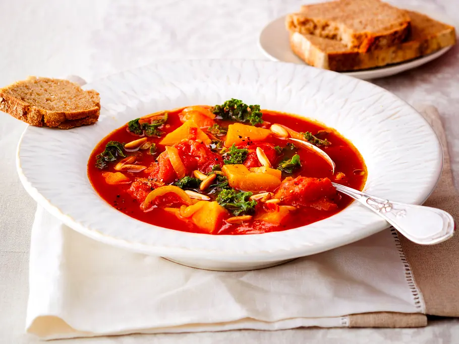 Kräftiger Eintopf mit Süßkartoffeln, Tomaten, Grünkohl und gerösteten Kernen in einem tiefen weißen Teller.