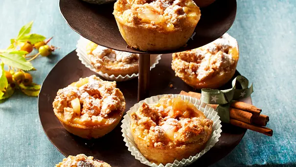 Apfel-Crumble Muffins mit Streuseln und Puderzucker liegen auf einer Etagere mit Zimtstangen. - Foto: LECKER @ Bauer Media Group