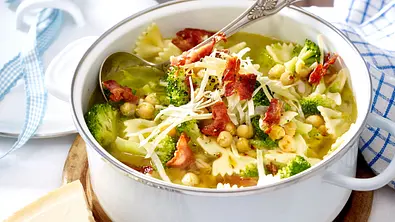 Schleifchennudelsuppe mit Brokkoli, Kichererbsen, knusprigem Speck und geriebenem Käse in einem weißen Topf auf einem Holzbrett serviert. - Foto: LECKER @ Bauer Media Group