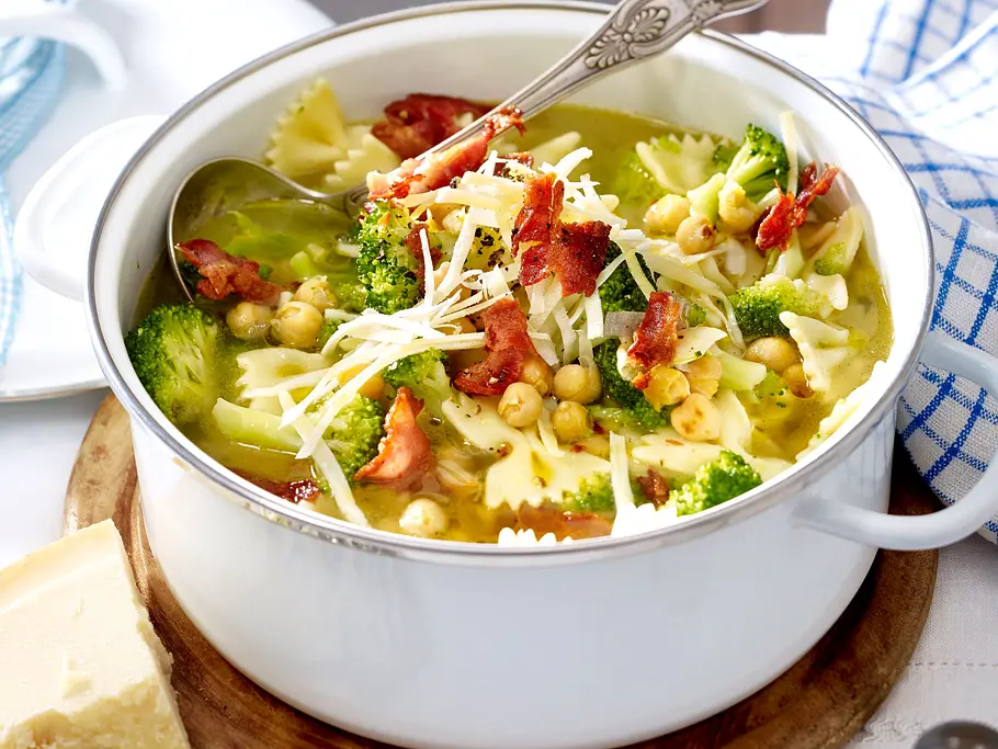 Schleifchen-Nudelsuppe mit Brokkoli und Kichererbsen Rezept Schleifchennudelsuppe mit Brokkoli, Kichererbsen, knusprigem Speck und geriebenem Käse in einem weißen Topf auf einem Holzbrett serviert.