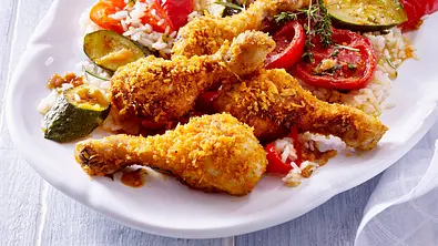 Panierte Hähnchenkeulen liegen mit Reis, Zucchini, roter Paprika, Tomaten und Kräutern auf einer weißen Servierplatte. - Foto: LECKER @ Bauer Media Group