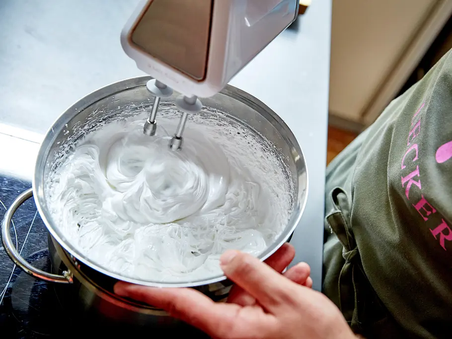 Mit einem Handmixer wird Eiweiß in einer Metallschüssel steif geschlagen, daneben steht eine Person mit Schürze.