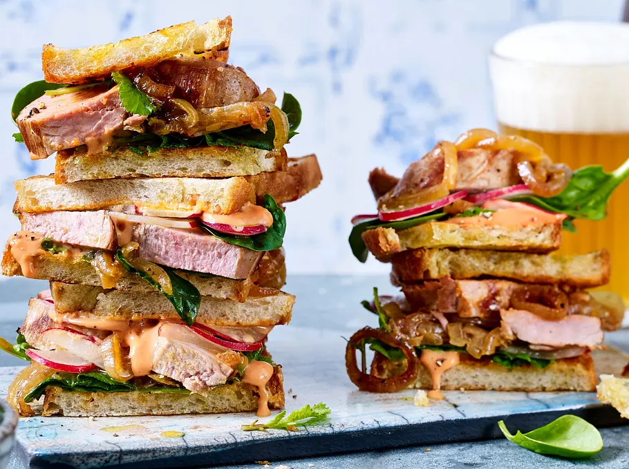 Mehrfach gestapelte Sandwiches aus geröstetem Brot, gefüllt mit saftigem Schweinefilet, Rucola, Radieschen und cremiger Soße, angerichtet auf einem hellen Schneidebrett auf grauer Steinplatte.
