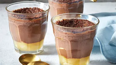 Drei Gläser mit gelb-braun geschichtetem Dessert und dunklen Bröseln stehen mit goldfarbenen Löffeln auf einem hellen Tisch. - Foto: LECKER @ Bauer Media Group