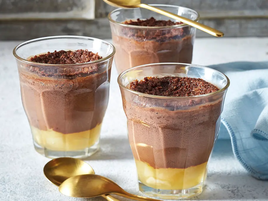 Mousse au chocolat mit Birnen Drei Gläser mit gelb-braun geschichtetem Dessert und dunklen Bröseln stehen mit goldfarbenen Löffeln auf einem hellen Tisch.