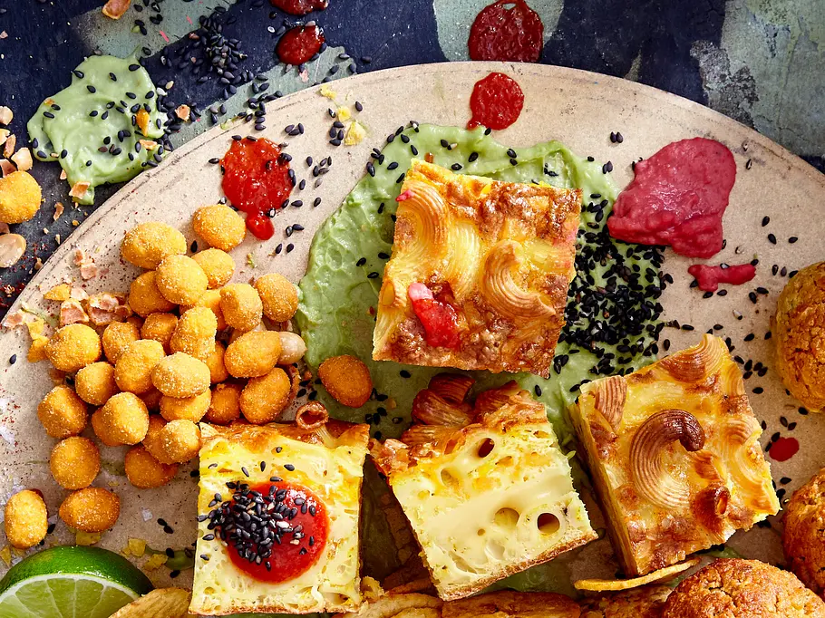 Mehrere goldbraune Mac-'n'-Cheese-Würfel liegen auf einer Snackplatte mit Dips, Nüssen, Limetten.