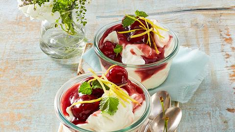 Glasschale gefüllt mit einer weißen Zitronencreme, welche mit einer roten Kirschsoße getoppt ist. Verziert ist es außerdem mit ganzen roten Kirschen, Zitronenschale und ein paar Minzblättern - Foto: House of Food / Bauer Food Experts KG
