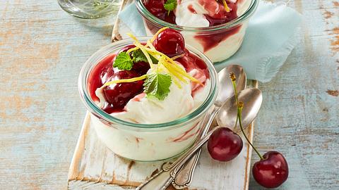 Glasschale gefüllt mit einer weißen Zitronencreme, welche mit einer roten Kirschsoße getoppt ist. Verziert ist es außerdem mit ganzen roten Kirschen, Zitronenschale und ein paar Minzblättern - Foto: House of Food / Bauer Food Experts KG