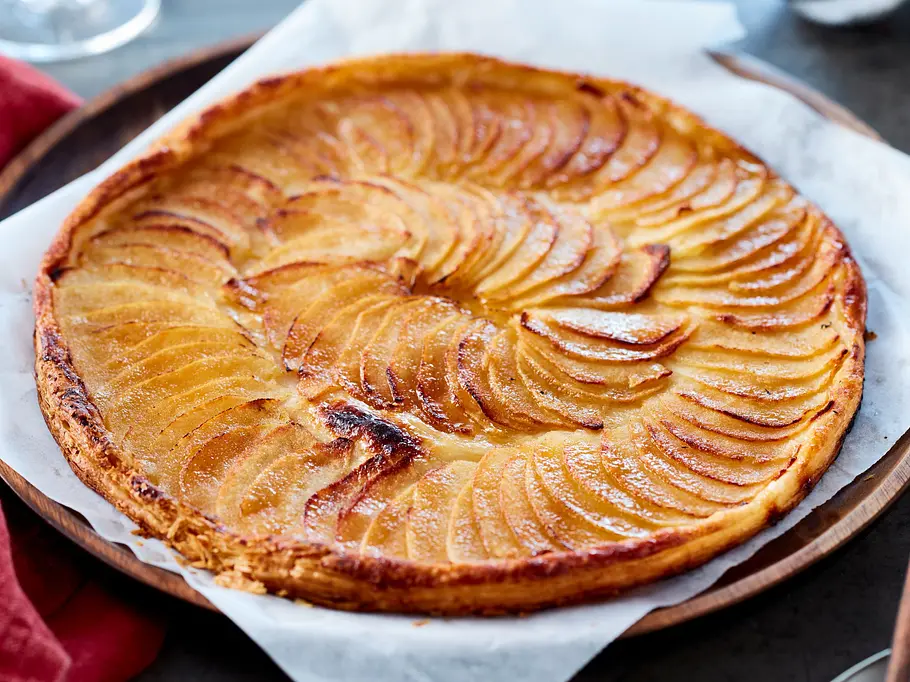 Dünne Tarte mit goldbraun gebackenen, fächerförmig gelegten Apfelscheiben auf buttrigem Teig, serviert auf Backpapier auf einem runden Holzbrett.
