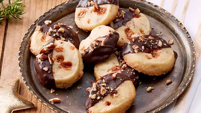 Runde Toffee-Pekan-Plätzchen, zur Hälfte in Zartbitterschokolade getaucht und mit gehackten Pekannüssen bestreut, serviert auf einem antik wirkenden Metallteller. - Foto: LECKER @ Bauer Media Group